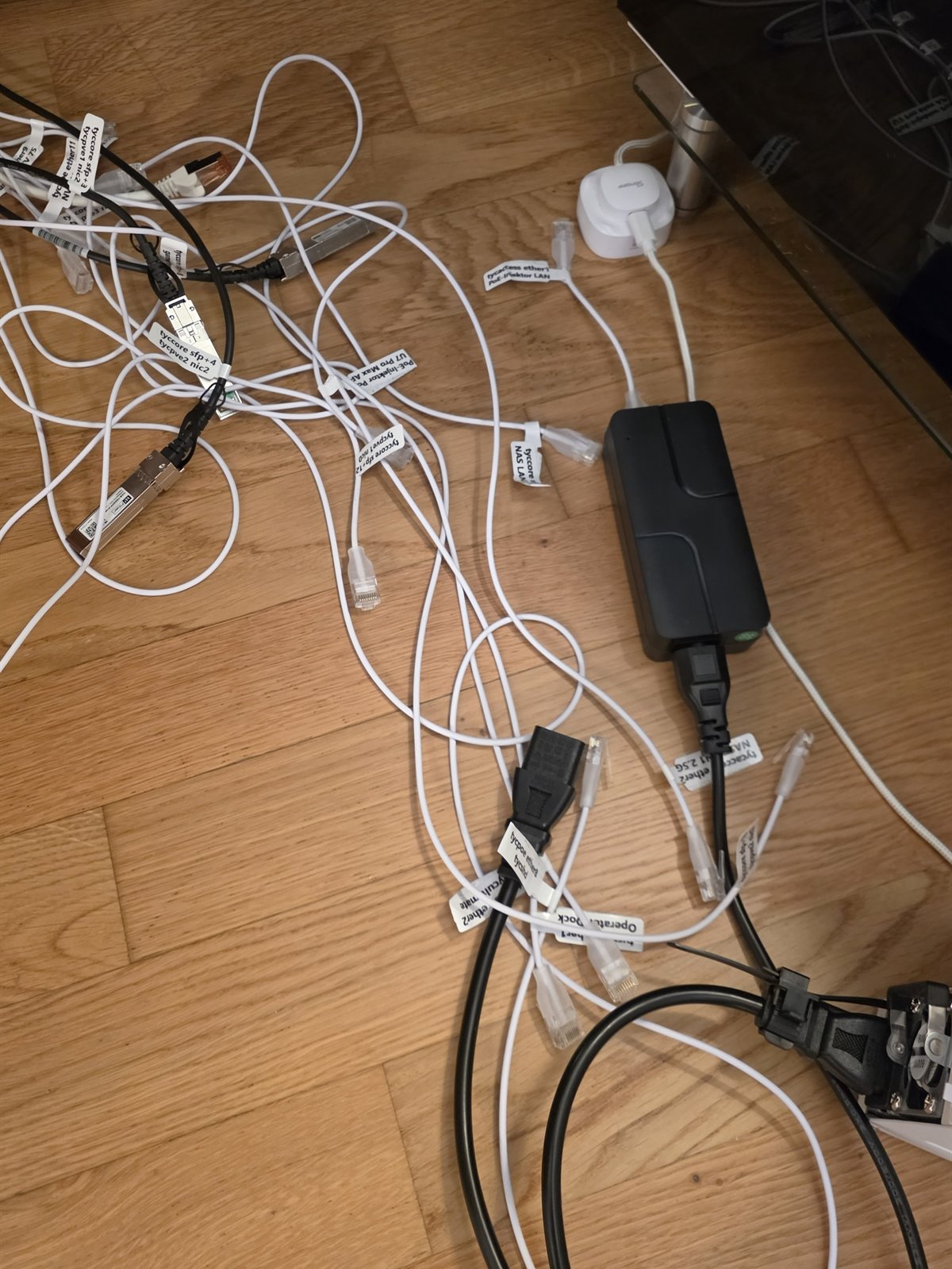 Labelled cables laid out on a wooden floor: white network cables and two black direct-attach cables, each with a white printed flag near the plug naming a switch port and the device at the far end, beside a black power brick.
