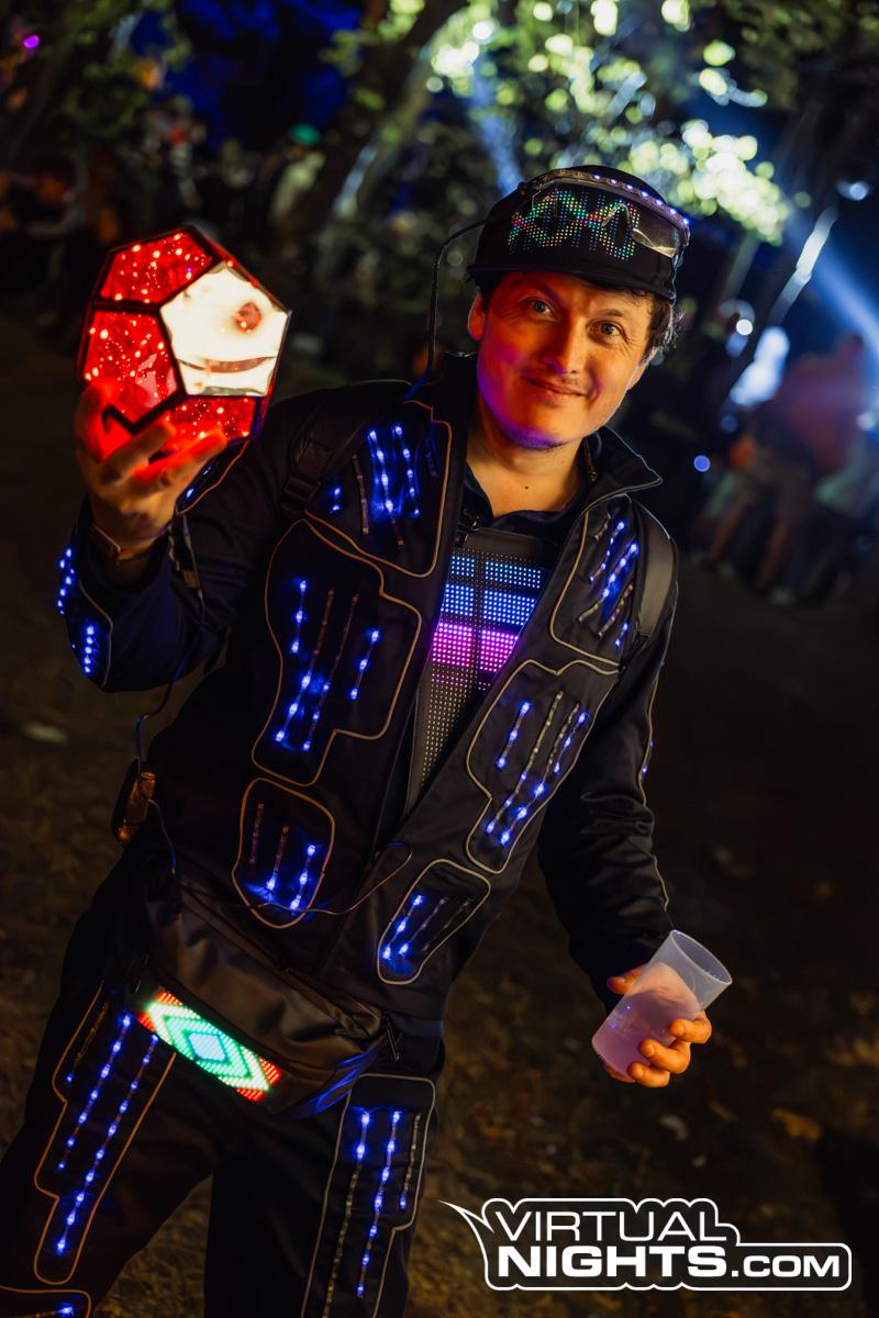 Me at Nibirii at night, in a black jacket lined with blue LED strips and a pixel panel on the chest, a light-up cap and glasses, holding up a glowing red dodecahedron lamp and a plastic cup.