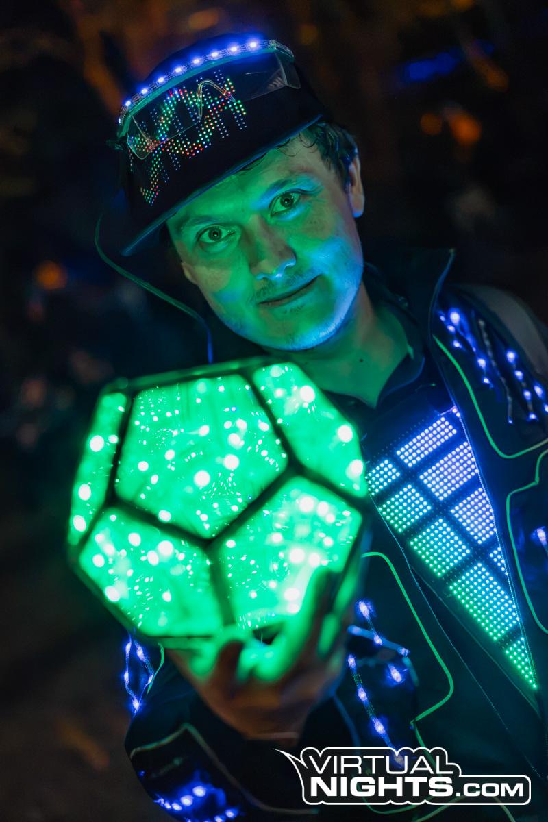 Me up close in green light, holding a glowing green dodecahedron lamp towards the camera; the LED jacket and the pixel panel light up blue and green, and text scrolls across the glasses.