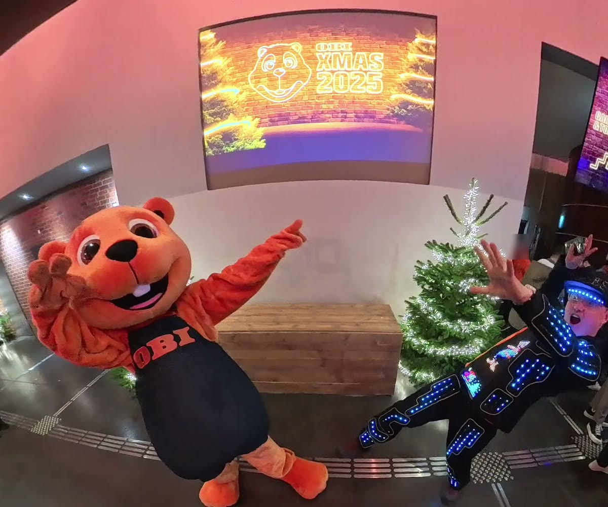 Me at the OBI Christmas party in the LED suit, one arm up and one leg kicked out, next to the OBI beaver mascot waving under a screen that reads XMAS 2025, a small lit Christmas tree between us.
