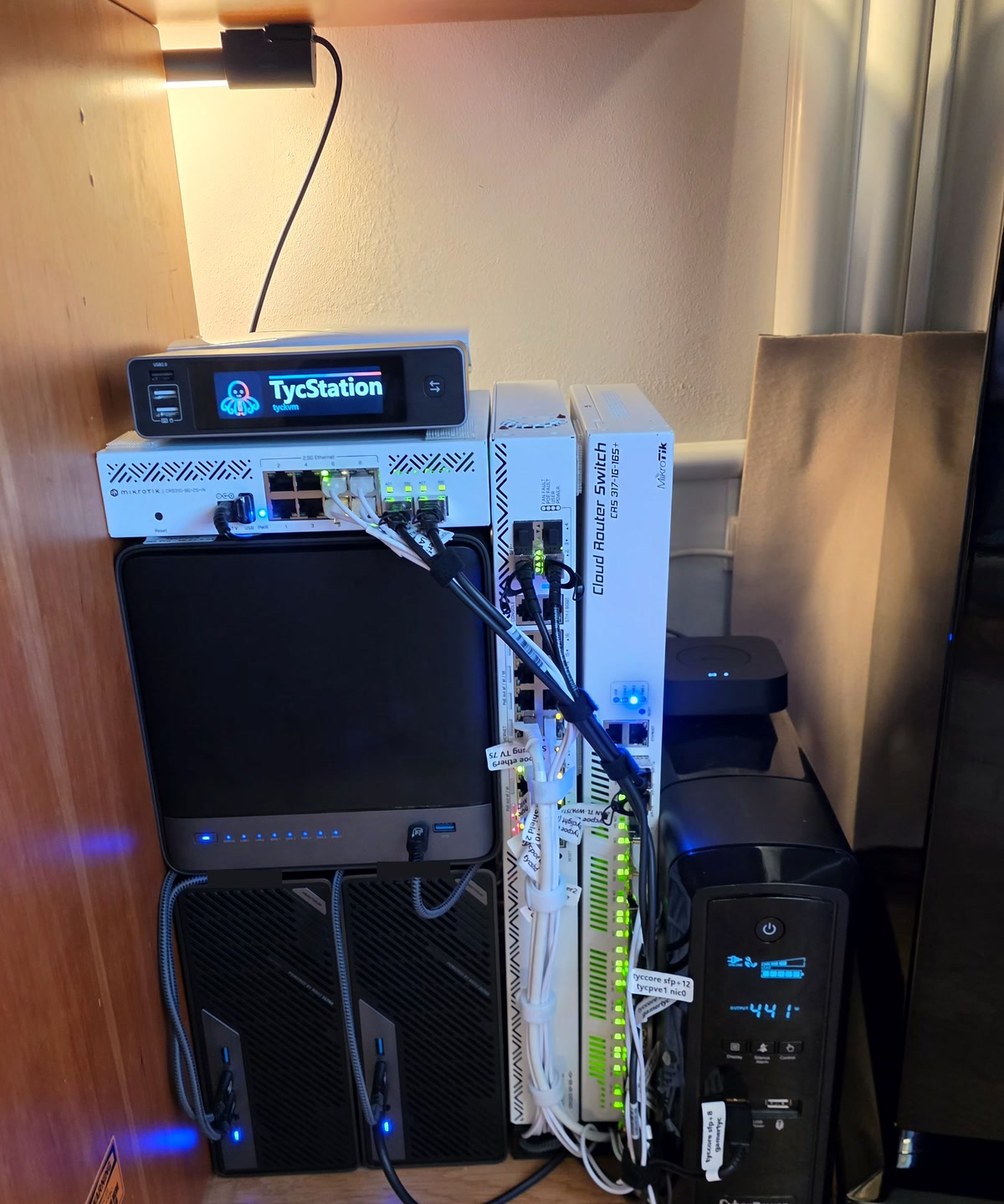 The rack on 20 September 2026, in the wooden alcove under the desk: a clip lamp under the shelf lighting the wall, the KVM with the station&rsquo;s name on its screen and the flat access switch on top of the black five-bay third node, the two MS-02 mini-PCs upright beneath it, the PoE switch and the core switch standing upright beside them with a bundled loom of white cables, the UPS on the right in front of the tall loudspeaker, and a white flag on every cable.