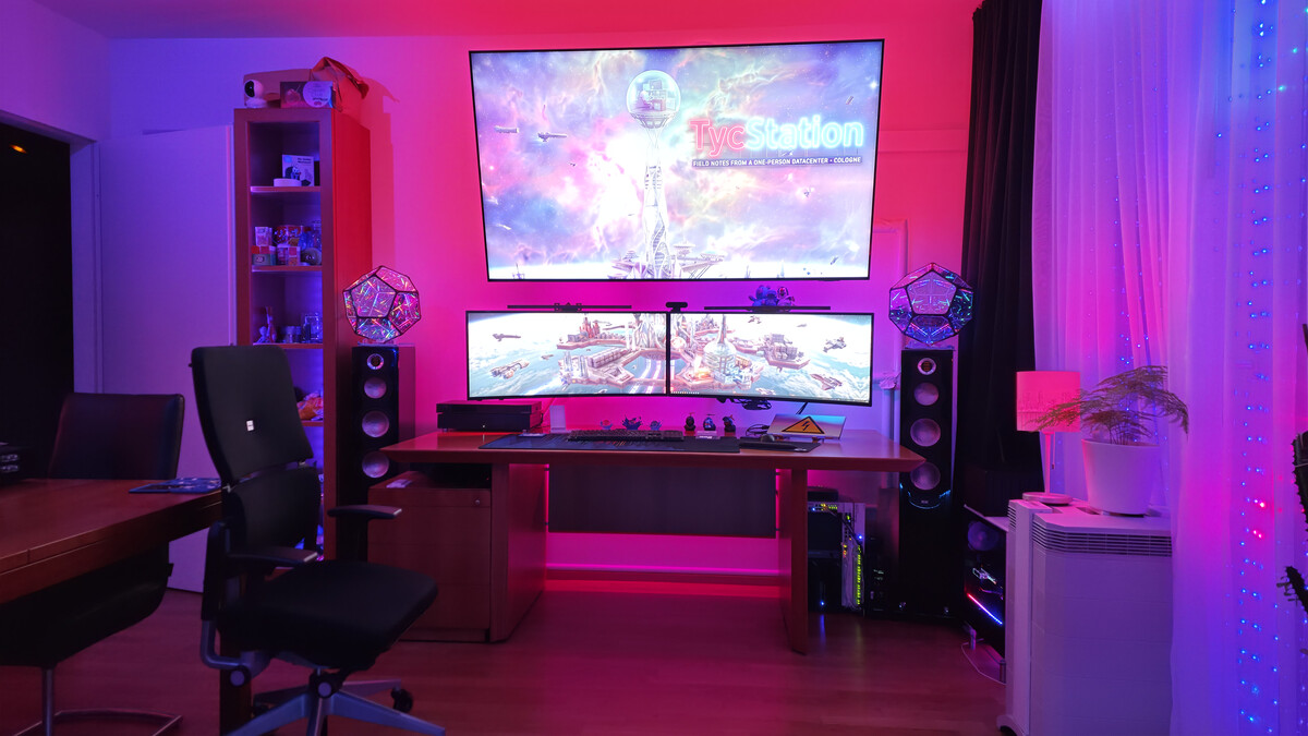 The room at night on 20 September 2026 in pink and blue light: a wooden desk and black chair beneath two curved monitors and a wall-mounted screen showing TycStation, two glowing dodecahedron lamps on the speakers, small computers under the desk, and a curtain of blue lights beside the window.