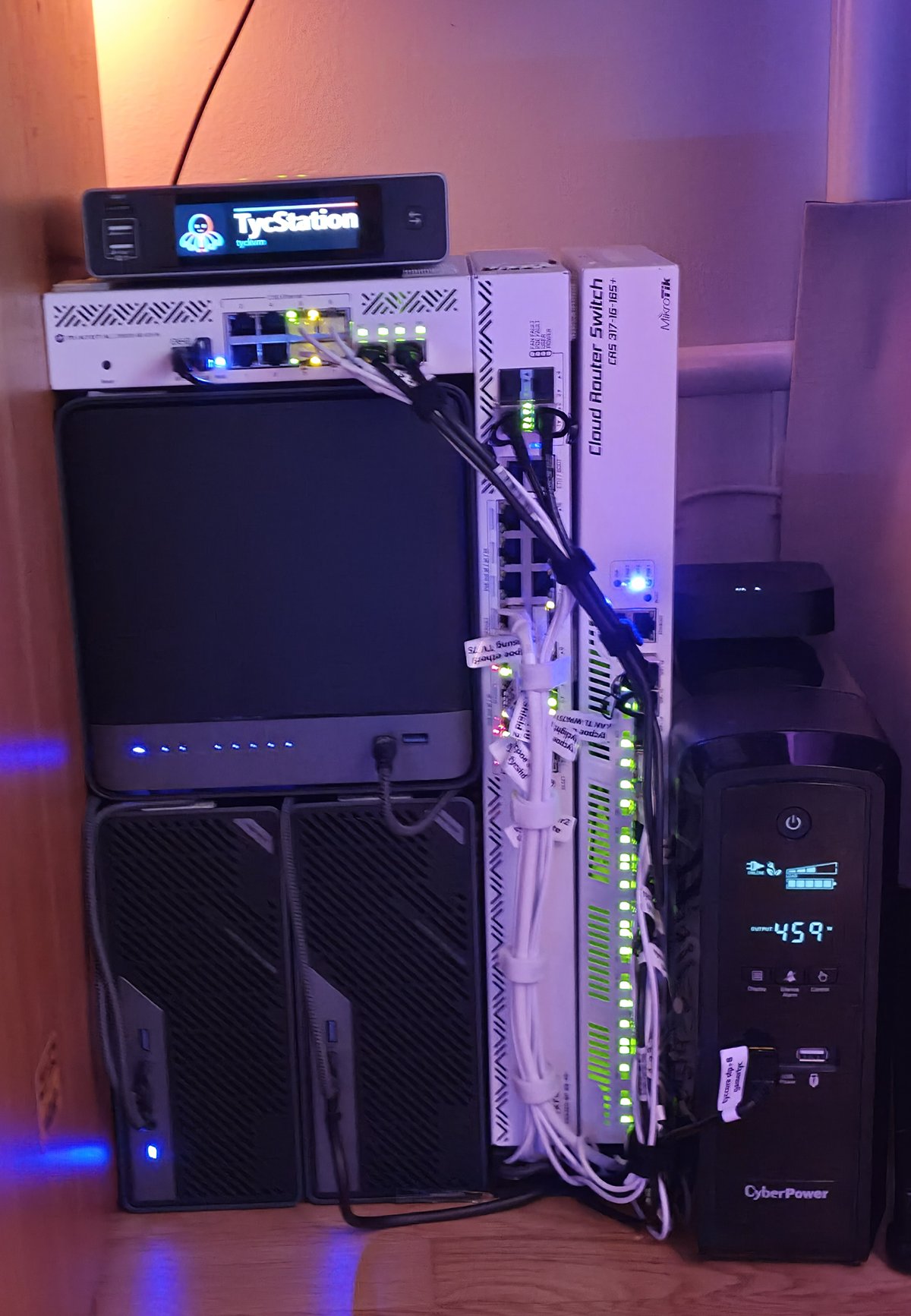 The stack under the desk at night, in warm lamp light and purple: a small screen showing the name TycStation and a flat switch on top of a black box with a row of blue lights, two small computers upright beneath it, two white switches standing upright beside them with rows of green lights and a bundle of white cables, and the battery on the right.