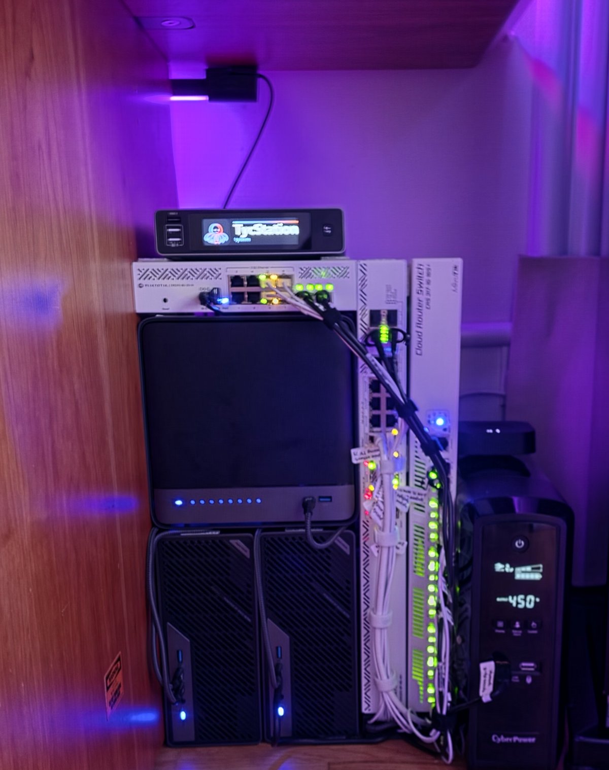 The stack under the desk at night, in purple light: a small screen showing the name TycStation and a flat switch on top of a black box with a row of blue lights, two small computers upright beneath it, two white switches standing upright beside them with rows of green lights and a bundle of white cables, and the battery on the right.